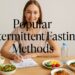 Smiling woman seated at a table with various healthy foods, a water bottle, and a fasting planner. Text overlay reads "Popular Intermittent Fasting Methods."